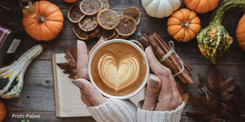 Person in a cozy white sweater holding a heart-shaped latte surrounded by autumn elements like pumpkins, cinnamon sticks, dried oranges, and fall leaves—evoking warmth, self-care, and seasonal well-being
