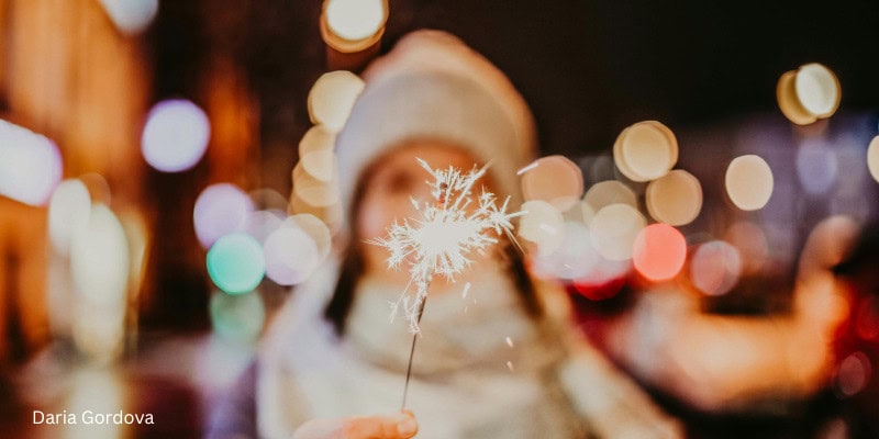 Person holding a lit sparkler on a winter city street with colorful bokeh lights in the background, wearing a hat and scarf, evoking a festive holiday atmosphere.