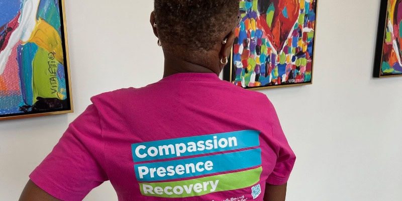 Person wearing a bright pink Cone Health “Compassion, Presence, Recovery” T‑shirt standing in front of colorful heart‑themed artwork in an indoor wellness space.