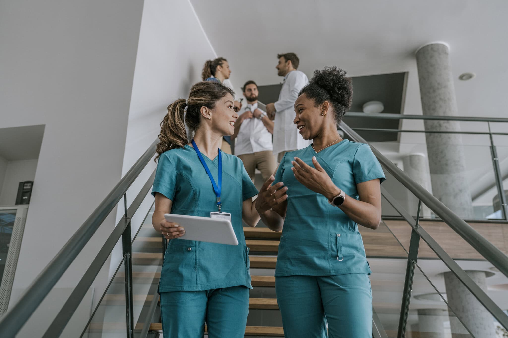 Two female doctors, wearing blue scrubs, are walking up a staircase in a hospital, chatting together. They appear to be in a good mood. There are other medical professionals walking in the background.