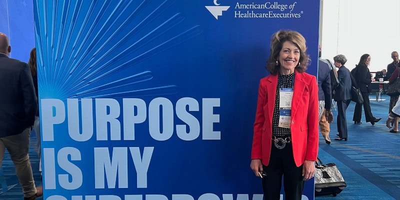 🖼️ Alt Text (SEO‑Optimized) Woman standing in front of a blue American College of Healthcare Executives display that reads “Purpose Is My Superpower” at a professional healthcare leadership event.