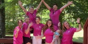 Group of Well‑Being Coaches wearing bright pink shirts posing together outdoors on a wooden deck, smiling and celebrating during a retreat.