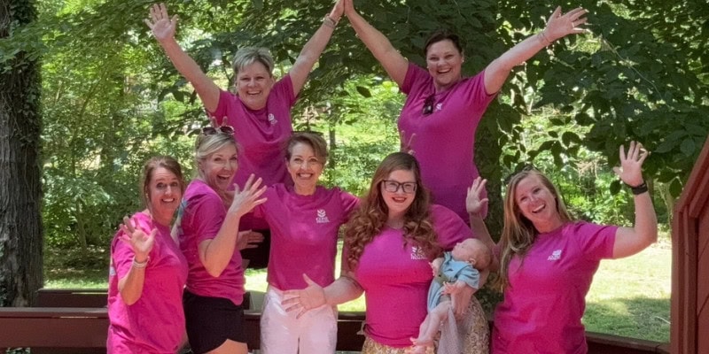 Group of Well‑Being Coaches wearing bright pink shirts posing together outdoors on a wooden deck, smiling and celebrating during a retreat.