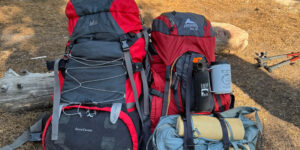 Two fully loaded hiking backpacks resting at a forest campsite with trekking poles and gear.